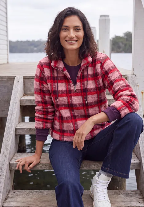 Vrouw zit ontspannen op houten treden, draagt rood geruit vest, jeans en witte sneakers aan het water.