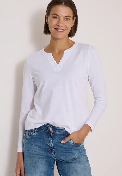 Frau in weißem Langarmshirt und Jeans, lächelnd, eine Hand in Tasche.