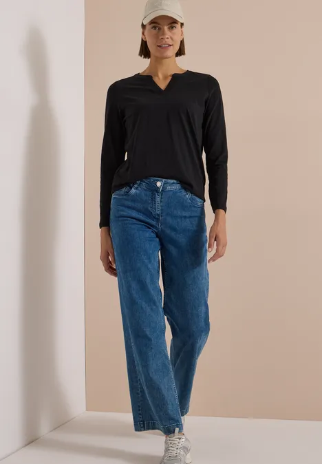 Langarm Shirt im Tunika-Look Black Frau trägt schwarzes Langarmshirt, weite blaue Jeans, weiße Sneaker und hellen Baseballcap im Raum