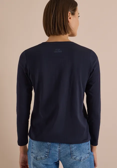 Rückenansicht einer Person in dunkelblauem Langarmshirt und Jeans vor beigem Hintergrund.
