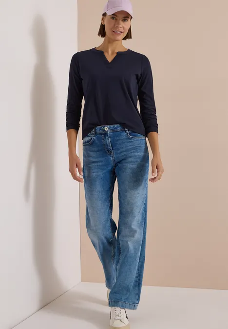 Frau mit dunkelblauem V-Shirt, weiten Jeans, weißen Sneakern und rosa Basecap vor neutraler Wand.