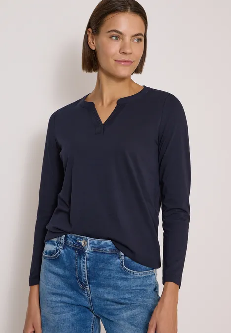 Frau mit kurzem Haar in langer, dunkelblauer Bluse und Jeans vor hellem Hintergrund.