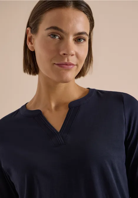 Frau mit kurzem braunem Haar trägt dunkelblaues Oberteil mit V-Ausschnitt, neutraler Hintergrund.