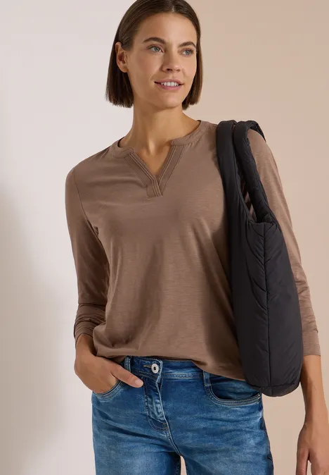 Frau mit braunem Langarmshirt, Jeans und schwarzer Schultertasche, lächelnd vor hellem Hintergrund.