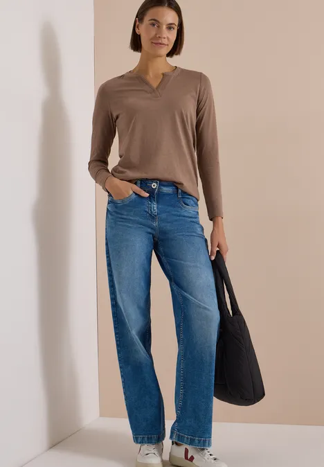 Frau mit braunem Langarmshirt, blauen Jeans, weißen Sneakern und schwarzer Tasche vor beigem Hintergrund.