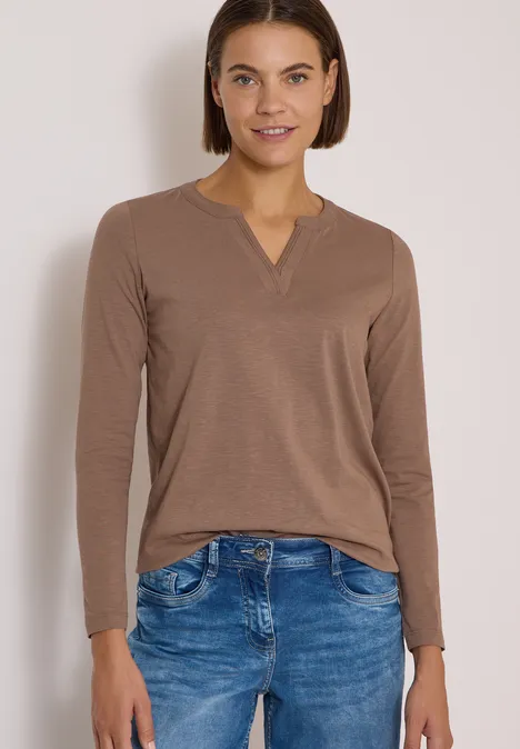 Frau in braunem Langarmshirt mit V-Ausschnitt und blauen Jeans vor hellem Hintergrund.