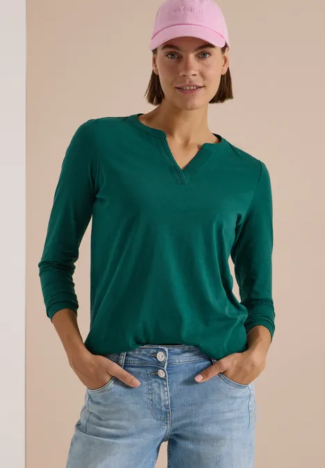 Langarm Shirt im Tunika-Look dark fir green Frau mit grünem V-Shirt, blauen Jeans und rosa Cap steht lässig mit Händen in Hosentaschen.