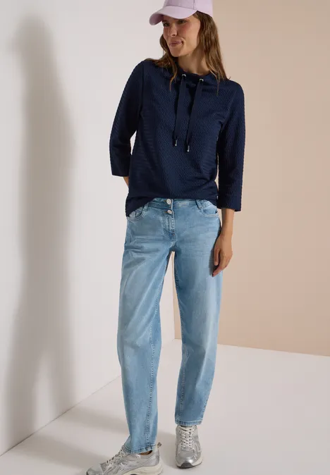 Frau in marineblauem Kapuzenpullover, hellblauen Jeans, grauen Sneakern und rosa Cap vor neutralem Hintergrund.
