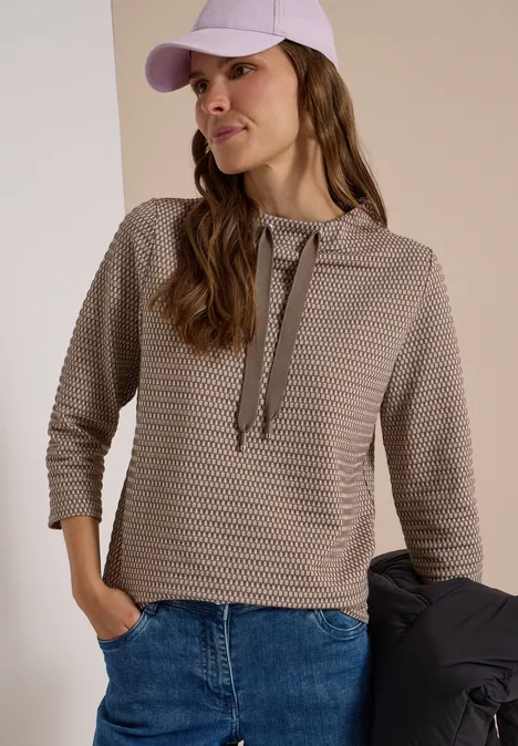 T-shirt à manches 3/4, col cheminée et texture urban taupe Femme portant un sweat à capuche texturé à manches 3/4, un jean et une casquette, tenant une veste.