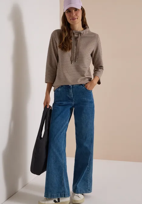 T-shirt à manches 3/4, col cheminée et texture urban taupe Femme en sweat à capuche rayé, casquette rose, jeans larges et grand sac noir sur fond clair.