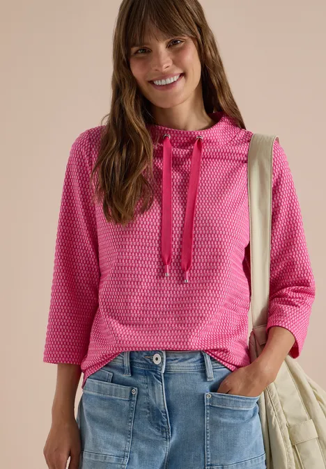 T-shirt à manches 3/4, col cheminée et texture pink flash Femme souriante en sweat à capuche rose et jean, sac sur l'épaule.