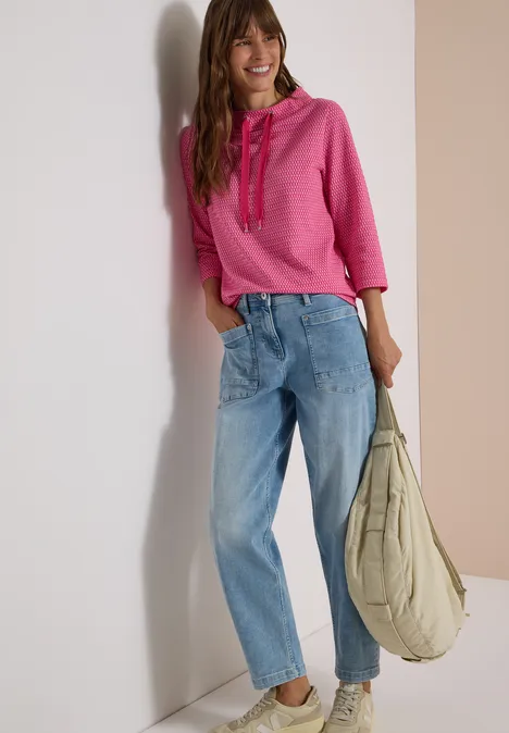 T-shirt à manches 3/4, col cheminée et texture pink flash Jeune femme souriante appuyée contre un mur, vêtue d’un sweat à capuche rose, d’un jean ample et portant un grand sac.