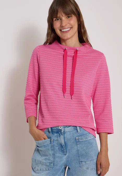 T-shirt à manches 3/4, col cheminée et texture pink flash Femme portant un sweat à capuche rose à motifs à manches 3/4 et un jean bleu ample, souriante.