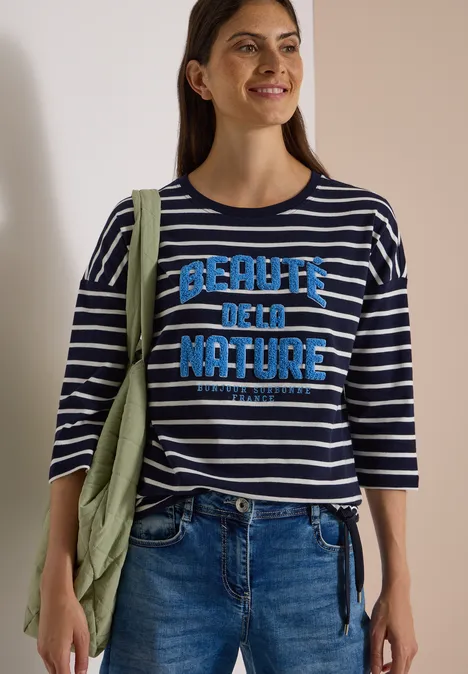 Femme en jean et marinière à rayures, slogan «Beauty of Nature» et veste verte sur l’épaule.