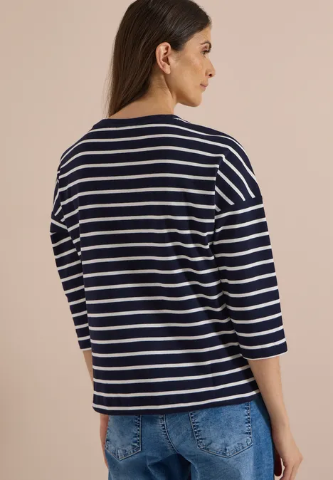 Femme de dos en marinière bleu marine et blanc à manches 3/4, portant un jean sur fond beige.