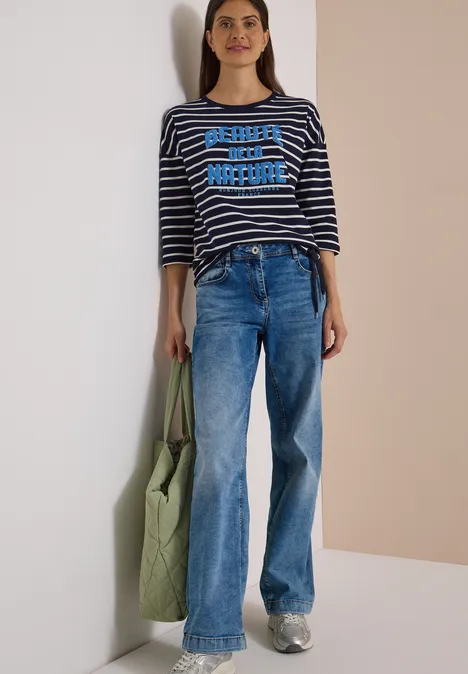Une femme en jean bleu, t-shirt rayé et sac vert appuyée de manière décontractée contre un mur clair.