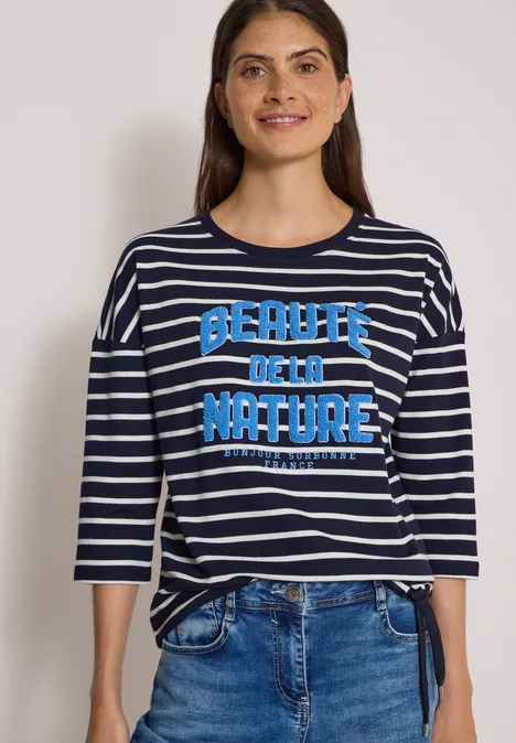 Une femme porte un t-shirt bleu à rayures blanches 'Beauty of the Nature' et un jean.
