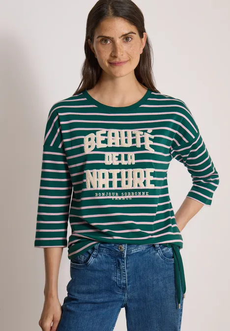 Eine lächelnde Frau trägt ein grünes, gestreiftes 3/4-Arm-Shirt mit Jeans.