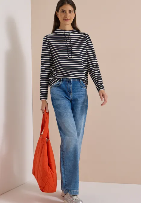 Frau in gestreiftem Pullover, weiten Jeans und orangefarbener Tasche vor heller Wand