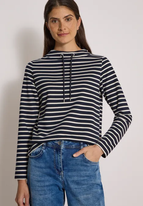 Frau mit blau-weiß gestreiftem Shirt und Jeans vor hellem Hintergrund.