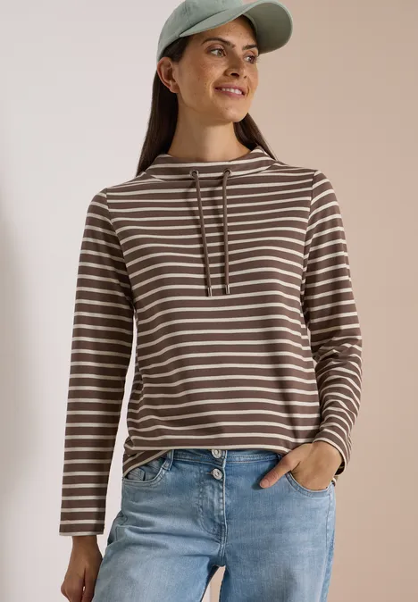 Frau mit gestreiftem Kapuzenshirt und Jeans, lächelt vor neutralem Hintergrund.