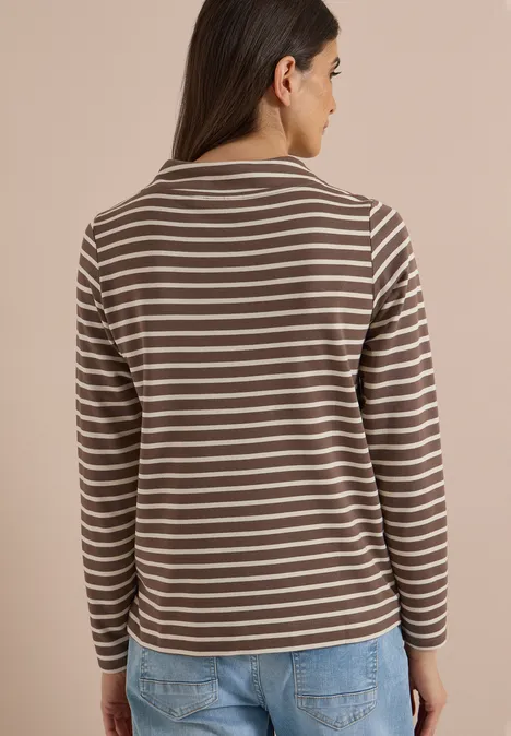 Frau von hinten in braun-weiß gestreiftem Langarmshirt und blauen Jeans vor hellem Hintergrund.