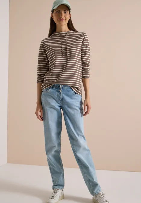 Frau in gestreiftem Sweater mit Kapuze, Jeans und Cap vor beigem Hintergrund.