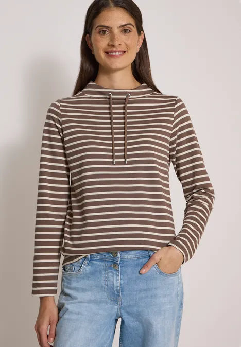 Junge Frau mit braun-weiß gestreiftem Longsleeve-Hoodie und Jeans, eine Hand in Tasche, lächelnd.
