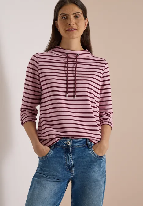 Vrouw in roze-zwart gestreepte capuchontrui en blauwe jeans, handen in zakken, neutrale achtergrond.