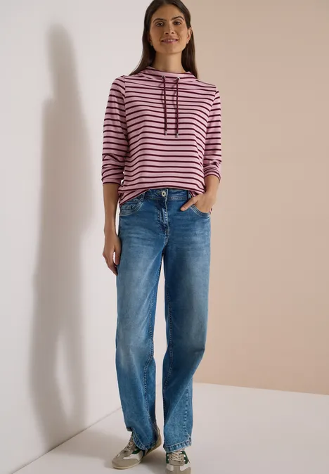 Vrouw in een roze-wit gestreepte trui met capuchon en losse blauwe jeans, staand voor een neutrale achtergrond.