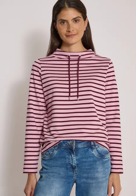 Jonge vrouw lacht in een roze-zwart gestreepte hoodie en blauwe jeans tegen een neutrale achtergrond.