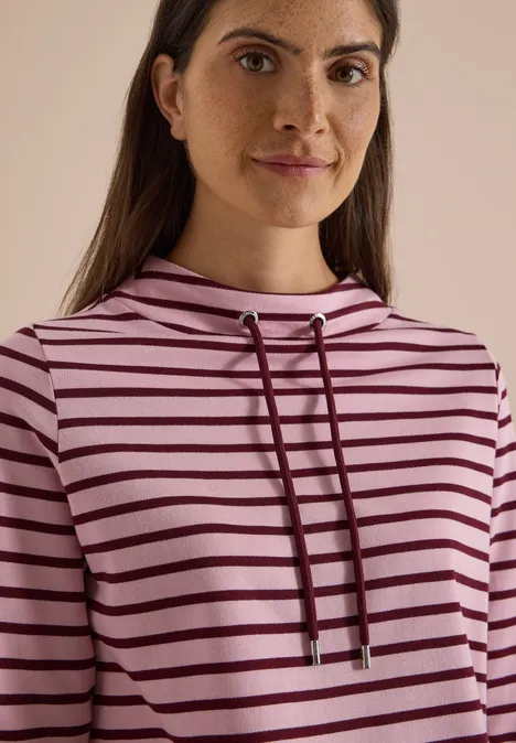 Vrouw met roze gestreepte capuchontrui en koordjes, donker haar en eenvoudige oorbellen.