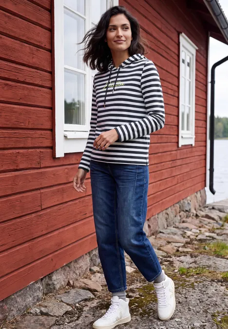 Shirt met lange mouwen, capuchon en strepen urban dark blue Frau in gestreiftem Pulli und Jeans spaziert vor einem roten Holzhaus am Wasser.