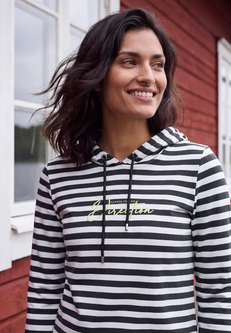 Shirt met lange mouwen, capuchon en strepen urban dark blue Frau im schwarz-weißen Hoodie mit gelbem Aufdruck lächelt vor roter Fassade.