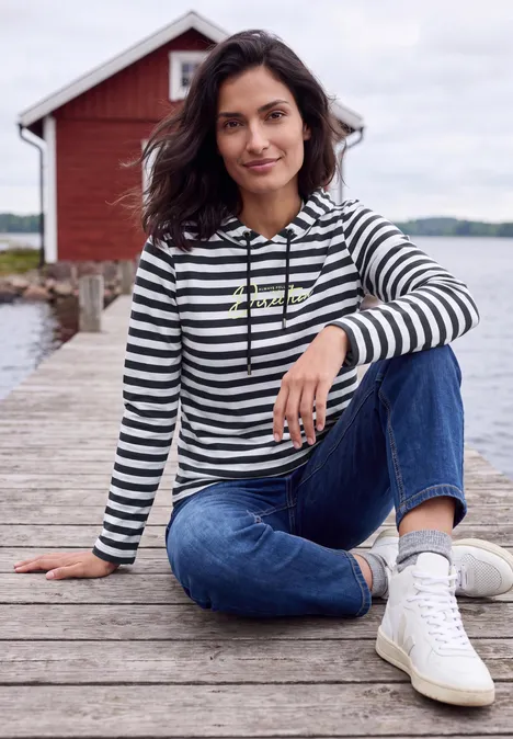 Shirt met lange mouwen, capuchon en strepen urban dark blue Frau in gestreiftem Hoodie und Jeans sitzt entspannt auf einem Steg am Wasser vor einem roten Bootshaus.