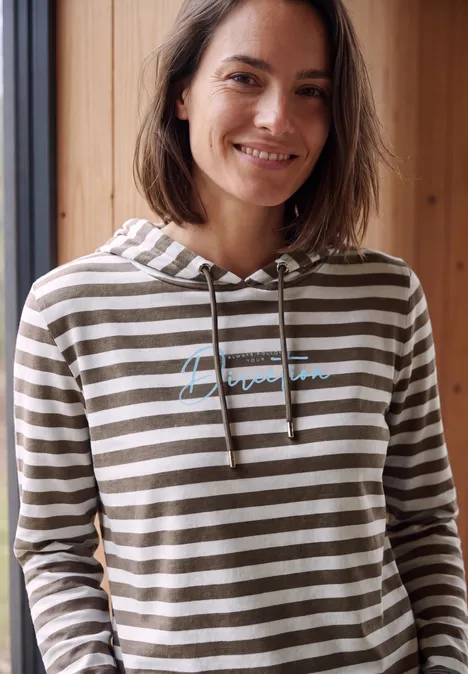 Eine lächelnde Frau in gestreiftem Hoodie vor einem Fenster und Holzpaneelen.