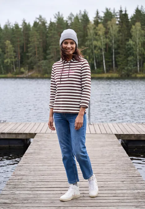Shirt met lange mouwen, capuchon en strepen red wine Eine lächelnde Frau in gestreiftem Pullover, Jeans und Mütze steht auf einem Steg vor einem See und Wald.