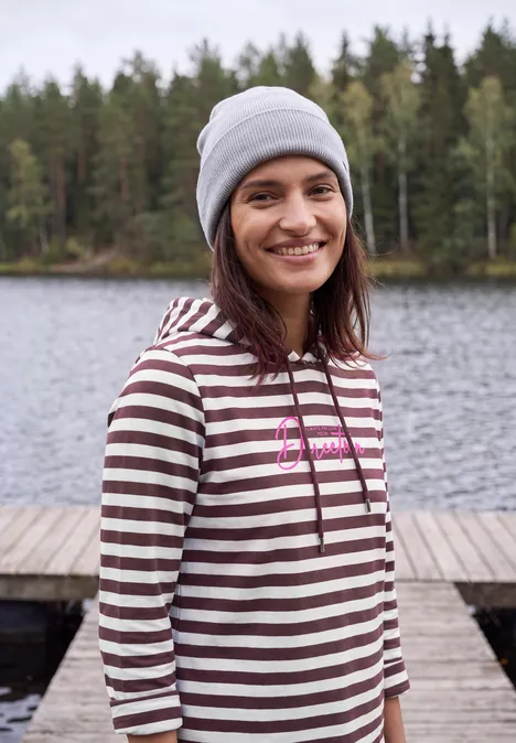 Shirt met lange mouwen, capuchon en strepen red wine Eine lächelnde Frau in gestreiftem Hoodie und Mütze steht an einem See vor einem Wald auf einem Steg.