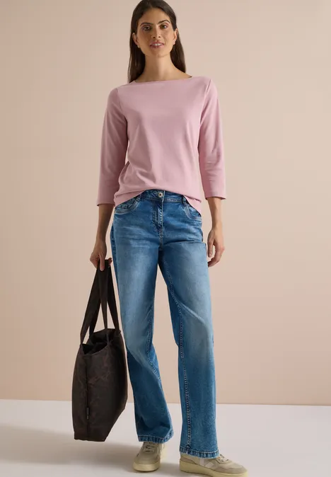 Eine Frau in rosa Dreiviertelarmshirt, blauen Jeans hält eine große, dunkle Tasche vor hellem Hintergrund.