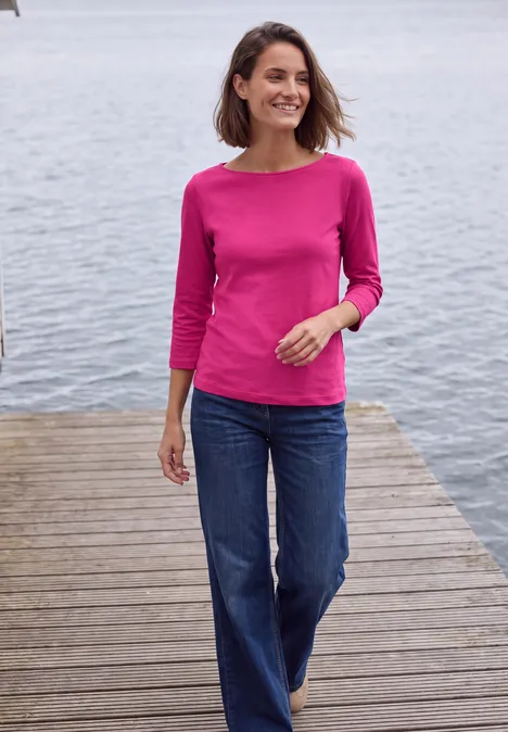 T-shirt basique à col bateau pink flash Frau in pinkem Shirt und Jeans spaziert auf einem Holzsteg am Wasser, lächelnd.