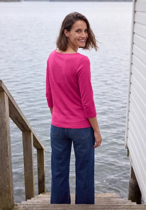 T-shirt basique à col bateau pink flash Eine lächelnde Frau in pinkem Pullover und Jeans steht auf Holztreppe am Wasser und blickt zurück.