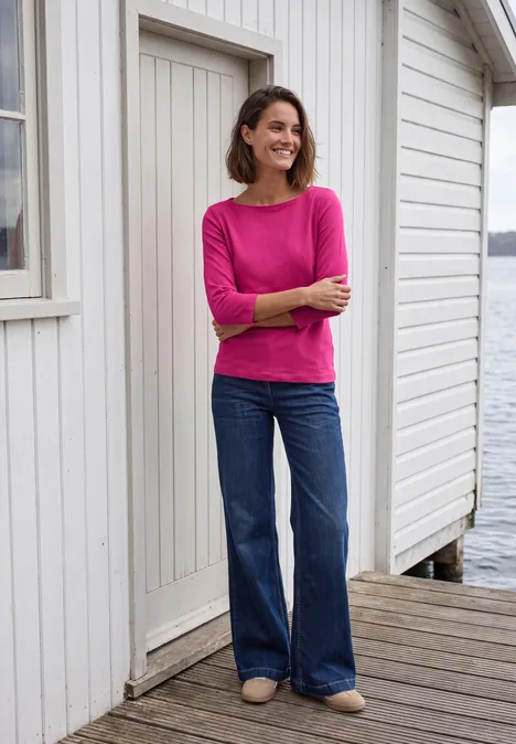 T-shirt basique à col bateau pink flash Frau lächelnd steht auf einem Steg vor weißem Haus am Wasser, trägt pinkes Shirt und weite Jeans.