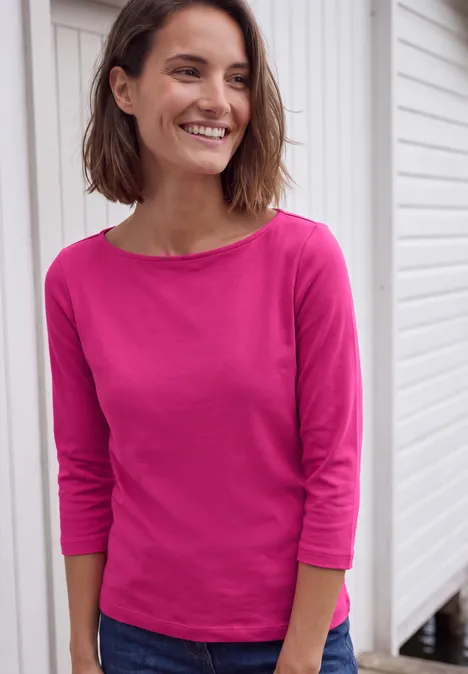 T-shirt basique à col bateau pink flash Eine lächelnde Frau in pinkem Oberteil vor weißer Fassade.
