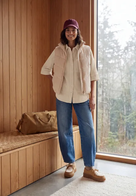 Femme souriante en doudoune beige, jean et casquette dans un intérieur en bois avec vue sur forêt.