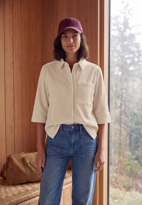 Une personne en jean, chemise et casquette se tient devant un mur en bois avec vue sur la nature.
