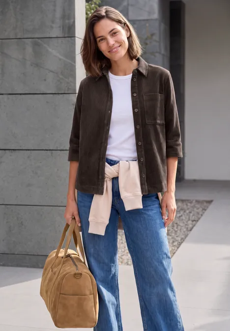 Blouse en jersey de velours côtelé à manches courtes, couleur unie truffle brown Frau in lässigem Outfit mit Jeans, weißem Shirt, brauner Jacke und Reisetasche vor moderner Fassade.