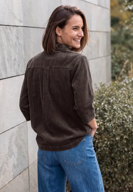 Blouse en jersey de velours côtelé à manches courtes, couleur unie truffle brown Eine Frau in brauner Cordjacke und blauer Jeans lehnt lächelnd an einer Steinmauer im Freien.