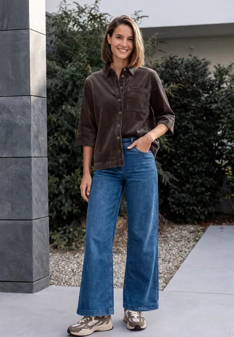 Blouse en jersey de velours côtelé à manches courtes, couleur unie truffle brown Eine Frau trägt braunes Cord-Hemd und blaue Schlagjeans, steht lächelnd vor einem Gebäude im Freien.
