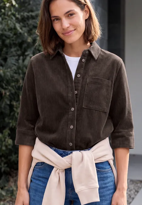 Blouse en jersey de velours côtelé à manches courtes, couleur unie truffle brown Lächelnde Frau in braunem Cordhemd, Jeans und hellbeiger Strickjacke um die Taille im Freien.