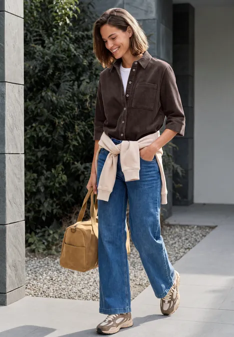 Blouse en jersey de velours côtelé à manches courtes, couleur unie truffle brown Lässig gekleidete Frau mit Tasche spaziert lächelnd an einem modernen Gebäude entlang.